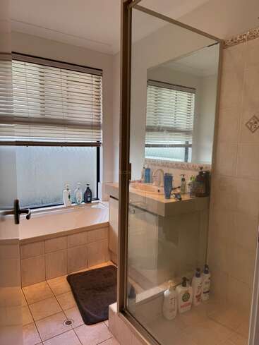 This bathroom features a glass shower, bathtub by a frosted window, tiled floor, beige walls, sink with toiletries, mirror, blinds, and a dark bath mat.