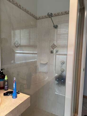 This image shows a tiled bathroom shower with a glass door. On the counter are toiletries, including a toothbrush, hair products, and pumps. Window blinds reflected.