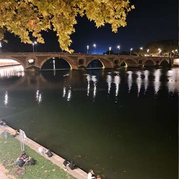 Uma cena noturna serena à beira do rio com uma ponte de pedra iluminada refletindo na água, folhas de outono acima e pequenos grupos de pessoas relaxando na margem do rio.