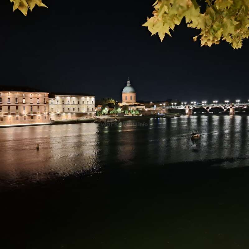 Uma cena noturna tranquila à beira do rio apresenta edifícios históricos iluminados, uma estrutura abobadada, uma ponte com luzes, reflexos na água e galhos frondosos pendentes.