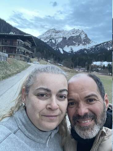 Ein Paar steht eng beieinander im Freien und lächelt, während im Hintergrund unter einem teilweise bewölkten Himmel malerische schneebedeckte Berge und ein rustikales Chalet zu sehen sind, was eine friedliche Atmosphäre schafft.