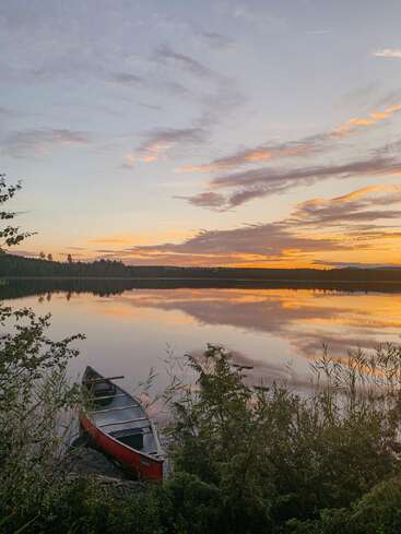 Un lago tranquilo al atardecer refleja las coloridas nubes. Una canoa roja descansa junto a la orilla, rodeada de follaje verde, creando una escena tranquila y natural.