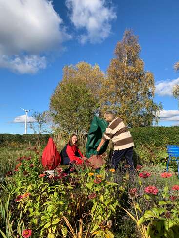 Zwei Menschen arbeiten an einem sonnigen Tag im Garten, umgeben von bunten Blumen und Herbstbäumen. Einer sitzt, der andere steht. Ein Windrad ist zu sehen.