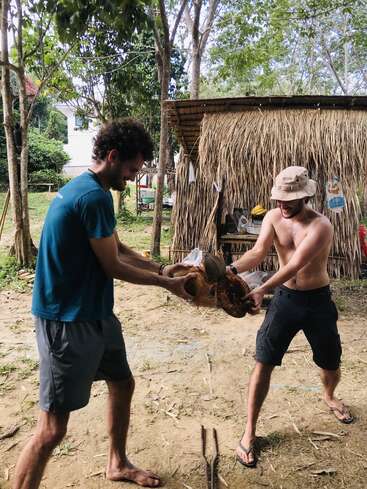Two men are outdoors, smiling and working together to open a coconut. One is shirtless, both look relaxed. Rustic hut and trees are in the background.
