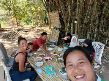 Un grupo de seis personas disfruta de una comida al aire libre en una mesa de bambú, rodeados de árboles y bambú. Todos sonríen, comparten comida, risas y un ambiente tranquilo.