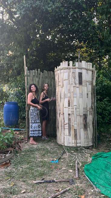 Zwei Frauen stehen draußen neben einer rustikalen Bambuskonstruktion, umgeben von viel Grün. Sie scheinen zu arbeiten oder etwas zu bauen, denn auf dem Boden liegen Werkzeuge und Materialien verstreut.