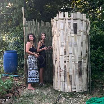 Deux femmes se tiennent debout à l'extérieur, près d'une construction rustique en bambou, au milieu de la verdure. Elles semblent être en train de travailler ou de construire, des outils et des matériaux étant éparpillés sur le sol.
