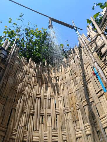 Dieses Bild zeigt eine Bambus-Außendusche mit einer Umzäunung aus geflochtenem Bambus, aus der das Wasser aus dem Duschkopf fließt, umgeben von Grün unter einem strahlend blauen Himmel.