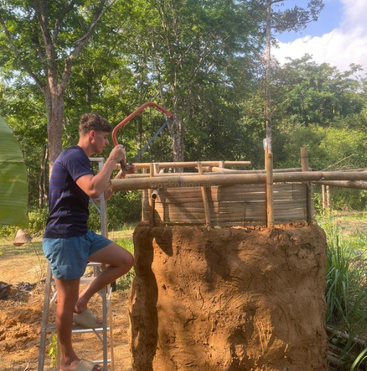 Ein Mann steht auf einer Leiter und sägt Bambus auf einem Bauwerk aus Lehm und Bambus, umgeben von üppiger grüner Vegetation.