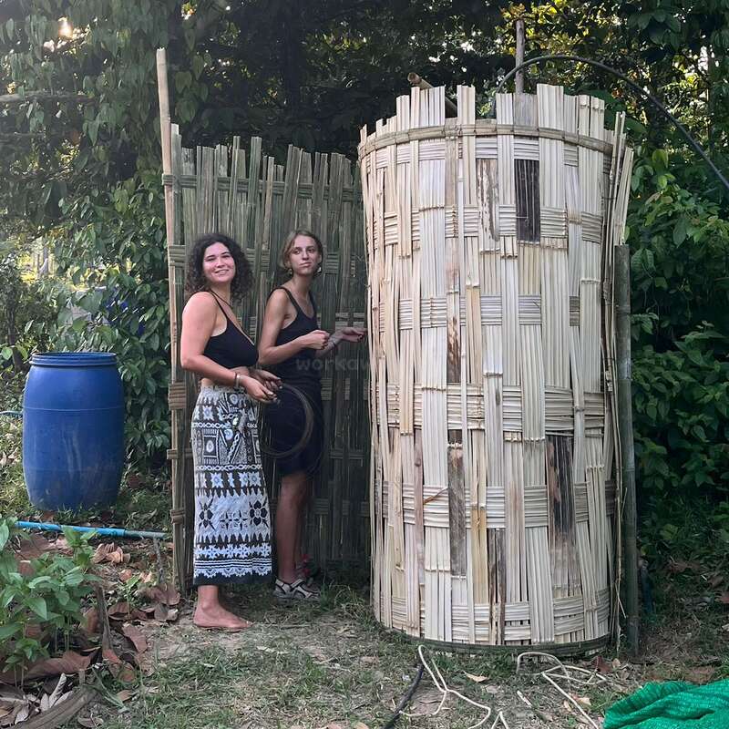 Zwei Frauen stehen draußen neben einer rustikalen Bambuskonstruktion, umgeben von viel Grün. Sie scheinen zu arbeiten oder etwas zu bauen, denn auf dem Boden liegen Werkzeuge und Materialien verstreut.