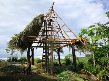 The image depicts a wooden structure with a thatched roof, under construction, situated on a tropical beach, surrounded by lush greenery and palm trees.