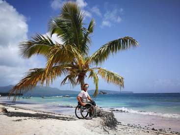The image depicts a man in a wheelchair sitting on a palm tree stump on a beach, with a serene ocean view and boats in the distance under a blue sky.