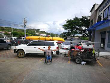 A man in a wheelchair is standing in a parking lot, gesturing with his arms outstretched, next to a white SUV and trailer with a yellow kayak on top.