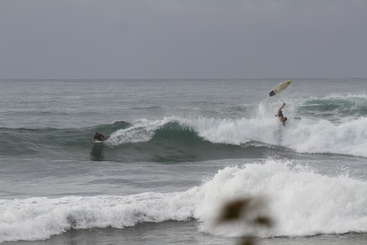 The image depicts a person falling from a wave while surfing, with the ocean and beach visible in the background. The scene is set against a natural, outdoor environment.
