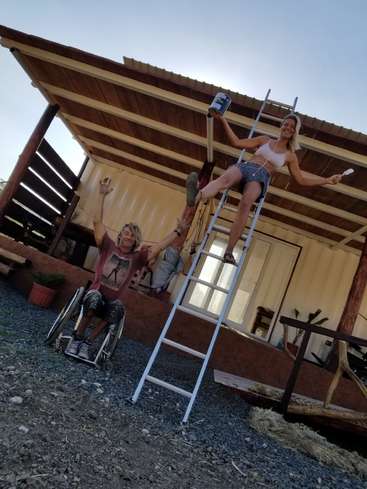 The image depicts a woman on a ladder, holding a paintbrush and can, with a man in a wheelchair beside her, standing in front of a house undergoing renovation.