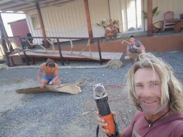 The image shows a man with long blonde hair holding a power tool, surrounded by people working on a wooden structure outside a building.