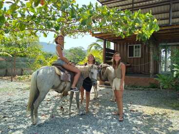 The image depicts three women posing with two white horses in front of a wooden building, set against a backdrop of trees and a mountainous landscape.
