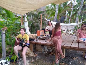The image depicts four individuals gathered in a tropical setting, surrounded by lush greenery, with two people playing guitars and two others seated on a wooden table.
