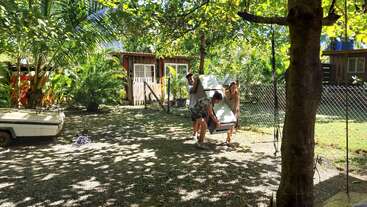 Three people are carrying a refrigerator through a sunlit, leafy backyard with small wooden houses, a fence, and a boat trailer under tropical trees and clear skies.
