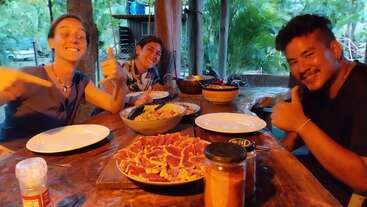 Three friends sit at a rustic wooden table, smiling and giving thumbs up, ready to enjoy a meal with dishes of meat, pasta, and condiments.