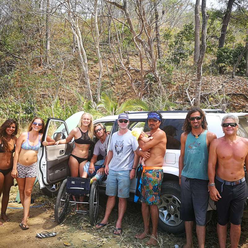 The image depicts a group of eight individuals, including two in wheelchairs, standing in front of a white SUV in a wooded area, posing for a photo.