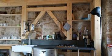 This cozy kitchen features wooden shelves, stone walls, utensils hanging above the sink, various jars and appliances, open shelving, a dishwasher, and a tall black floor lamp.