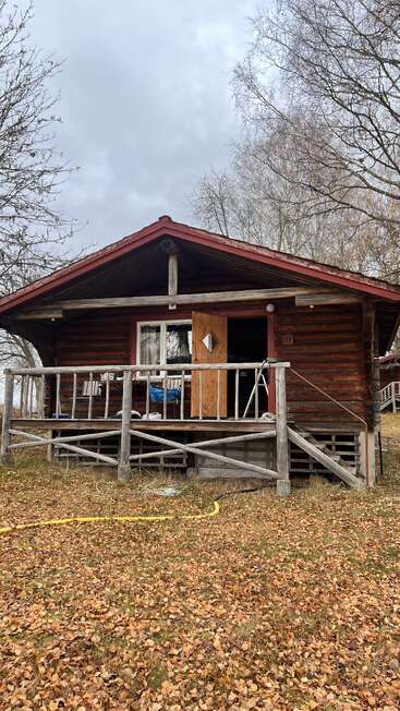 Une cabane rustique en bois avec un toit rouge se trouve au milieu d'arbres sans feuilles et de feuilles d'automne tombées au sol. La balustrade du porche est rugueuse et la porte d'entrée est ouverte.