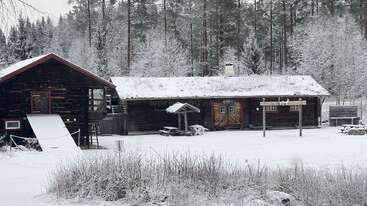 Uma cabana de madeira rústica e um celeiro estão em meio a uma paisagem de neve, cercados por árvores cobertas de gelo. Mesas de piquenique, placas e uma atmosfera serena de inverno completam a cena.