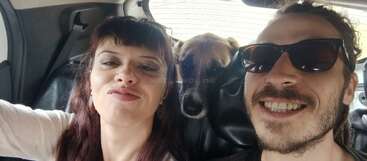A woman and a man take a selfie in a car, smiling happily. Between them, a brown dog looks curiously from the backseat, sharing the moment.