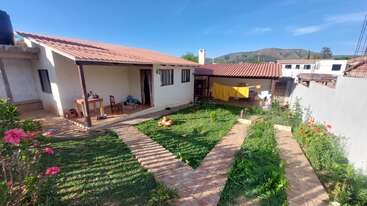 This image shows a cozy house with red tiled roof, a green yard, blooming flowers, a stone path, laundry drying, mountains, and blue sky.
