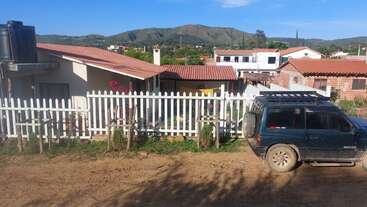 A rural scene with houses, red-tiled roofs, a white picket fence, parked black SUV, dirt road, green hills, clear sky, and garden plants.