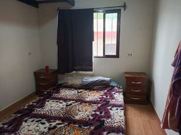 A simple bedroom with a double bed covered in a leopard print blanket, two wooden nightstands, a window with a dark curtain, and light-colored walls.