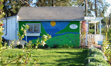 Um pequeno prédio com telhado inclinado exibe um mural colorido com colinas, céu e sol. Está rodeado por grama verdejante, flores e plantas de jardim.