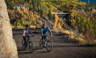 Deux personnes font du VTT sur un sentier de terre battue, entourées d'arbres aux couleurs de l'automne, avec en arrière-plan un pont en bois pittoresque et des collines au loin. Un cadre ensoleillé et paisible.