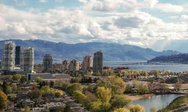 Un paysage urbain pittoresque composé de grands immeubles, d'arbres et d'un lac charmant, avec les montagnes en arrière-plan. Un pont enjambe le lac sous un ciel partiellement nuageux.