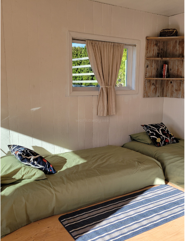 Une chambre chaleureuse et baignée de soleil, avec deux lits verts, des coussins colorés, un tapis rayé, des étagères en bois remplies de livres, des murs lambrissés de blanc et une fenêtre ornée de rideaux.