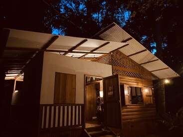 Una acogedora cabaña de madera se encuentra cálidamente iluminada por la noche, rodeada de altos árboles. El tejado ondulado y la acogedora puerta abierta crean un ambiente tranquilo y rústico.