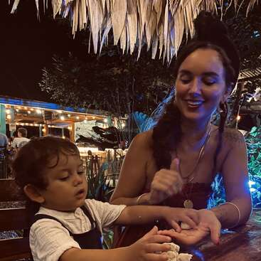 Una mujer y un niño pequeños se sientan juntos en un restaurante al aire libre por la noche, cálidamente iluminados por cuerdas de luces, sonriendo y jugando con algo en las manos.