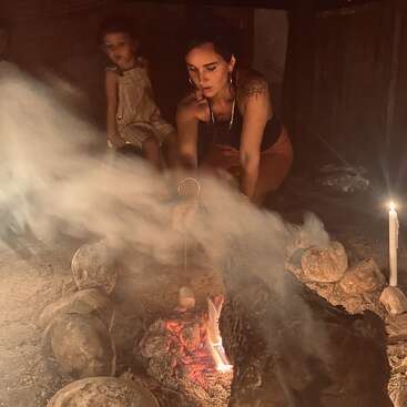 Une femme fait griller des marshmallows sur un feu de camp, entourée de pierres et de fumée. Un enfant est assis à proximité, tandis qu'une bougie allumée apporte une douce lumière supplémentaire.