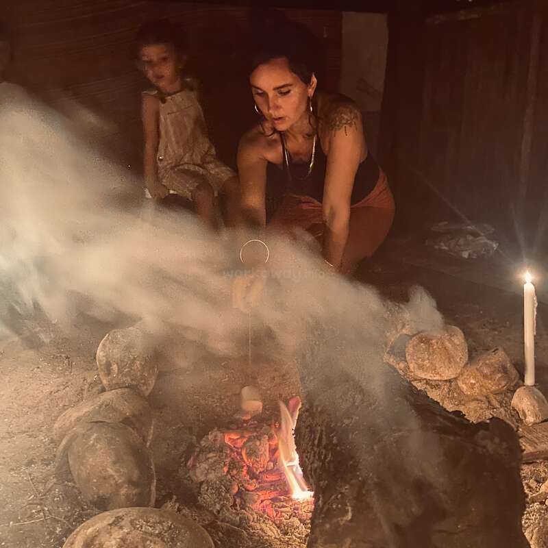 Uma mulher assa marshmallows em uma fogueira, cercada por pedras e fumaça. Uma criança senta-se por perto, enquanto uma vela acesa fornece uma luz adicional suave.