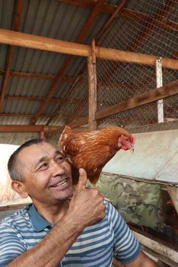 Un homme souriant, vêtu d'une chemise rayée, lève le pouce tandis qu'un poulet brun se perche sur son épaule à l'intérieur d'un poulailler rustique avec des poutres en bois.