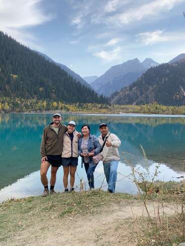 Quatre personnes sourient au bord d'un lac bleu serein, entouré de forêts de pins luxuriantes et de montagnes imposantes, sous un ciel partiellement nuageux, profitant d'un moment pittoresque en plein air.