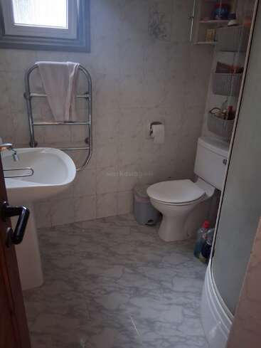 This bathroom features tiled floors and walls, a sink, toilet, towel rack, small window, shower with glass door, storage shelves, and assorted toiletries and cleaning products.