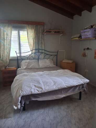 A cozy bedroom with a metal-framed bed, white bedding, two wooden nightstands, floral curtains, marble-patterned floor, and wooden shelves holding various items. Natural light streams in.