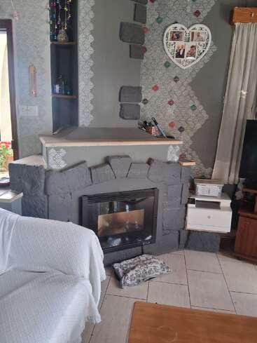 Cozy living room with a stone fireplace, heart-shaped photo frame, patterned walls, white couch, wooden furniture, small shelves, and a pillow on tiled floor.