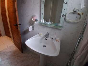 A small bathroom with a pedestal sink, a decorative mirror, toiletries, a hand towel holder, wooden door, tiled walls, and a rubber duck on the sink.