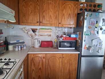 This cozy kitchen features wooden cabinets, a microwave, fridge with magnets, kettle, bread box, spice jars, gas stove, wine bottle, and various organized utensils.