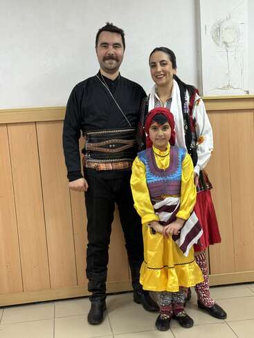 Ein Mann, eine Frau und ein Kind in farbenfroher Tracht posieren gemeinsam in einem Innenraum; sie stehen lächelnd vor einer Holzwand und einem weißen Hintergrund.