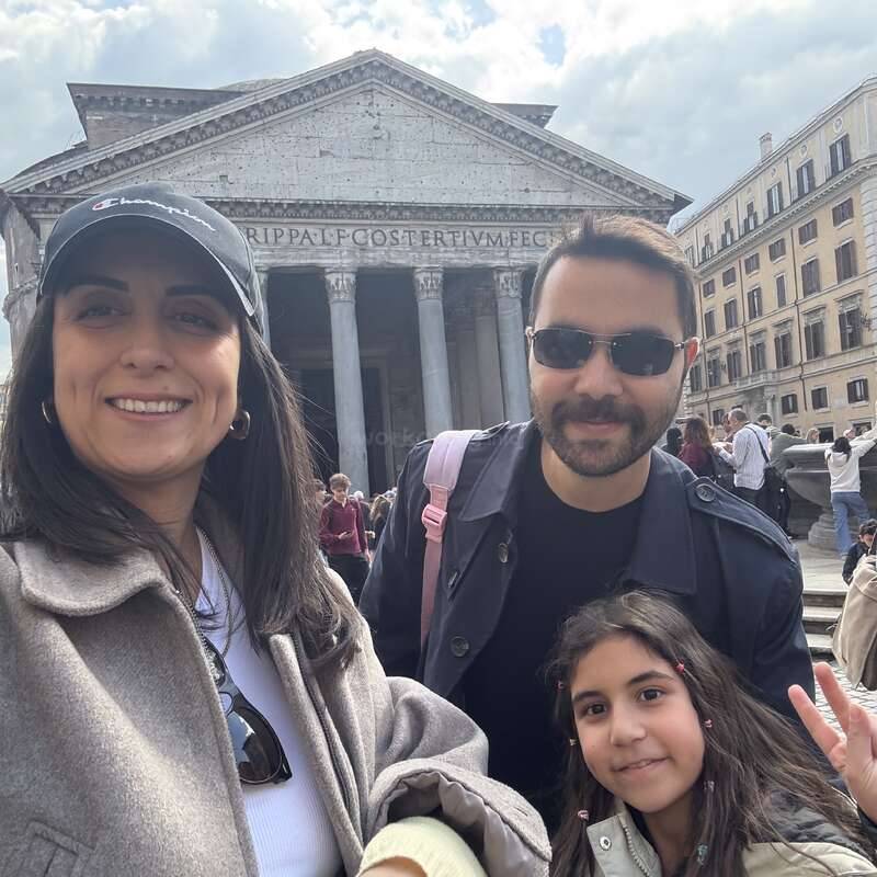 Eine lächelnde Frau, ein Mann und ein junges Mädchen machen ein Selfie vor dem Pantheon in Rom. Der Himmel ist bewölkt, und man sieht andere Touristen.