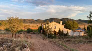 A rustic country house sits amidst dry grass and trees, with a dirt path leading to it. Rolling hills and cloudy skies complete the peaceful landscape.